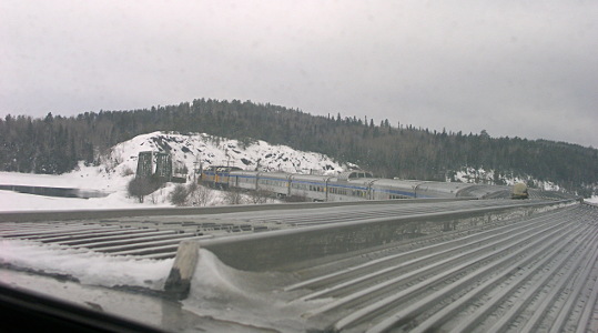 [The train is on a curved section of track getting ready to cross a bridge. Even though this view is from the last car, because the train is curving, the side view of the yellow engine and the first four silver cars are visible. The view of the remaining cars is just the roofs of them. The roof of the car immediately in front of the window is corrugated. There are tree covered hills beyond the bridge and snow covers the ground.]